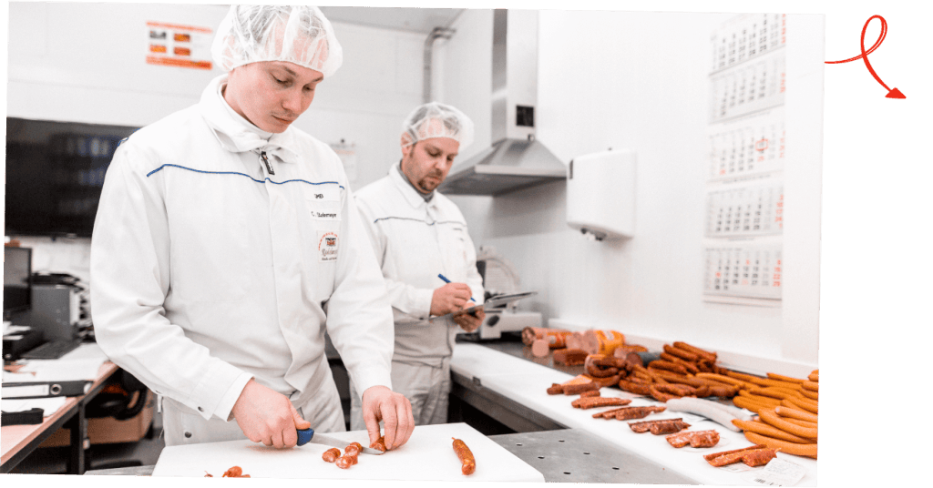 Zwei Mitarbeiter in weißer Arbeitskleidung und Haarnetz schneiden und kontrollieren Würste in der Produktion.