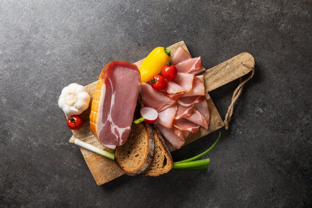 Rohschinken am Stück und in Scheiben, umgeben von Brotscheiben, Knoblauch, Paprika, Tomaten und Radieschen auf dunklem Untergrund.