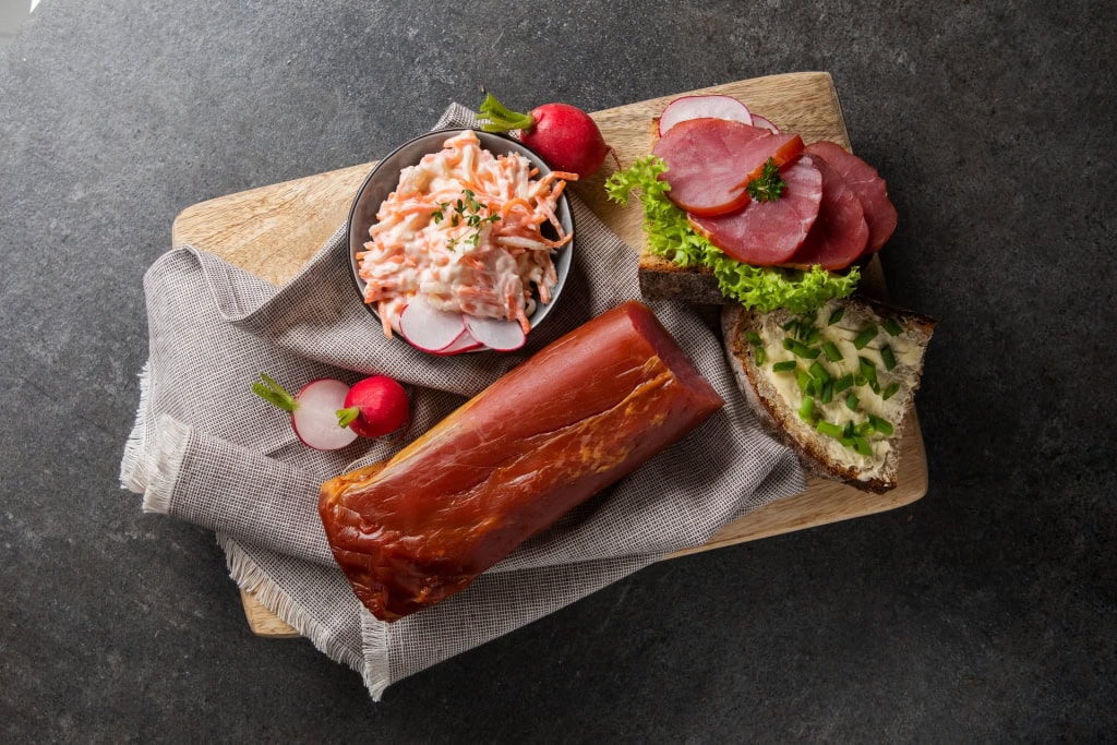 Geräuchertes Schinkenstück neben Krautsalat, Radieschen, einem Schinkenbrot mit Salat und einem Butterbrot mit Schnittlauch.