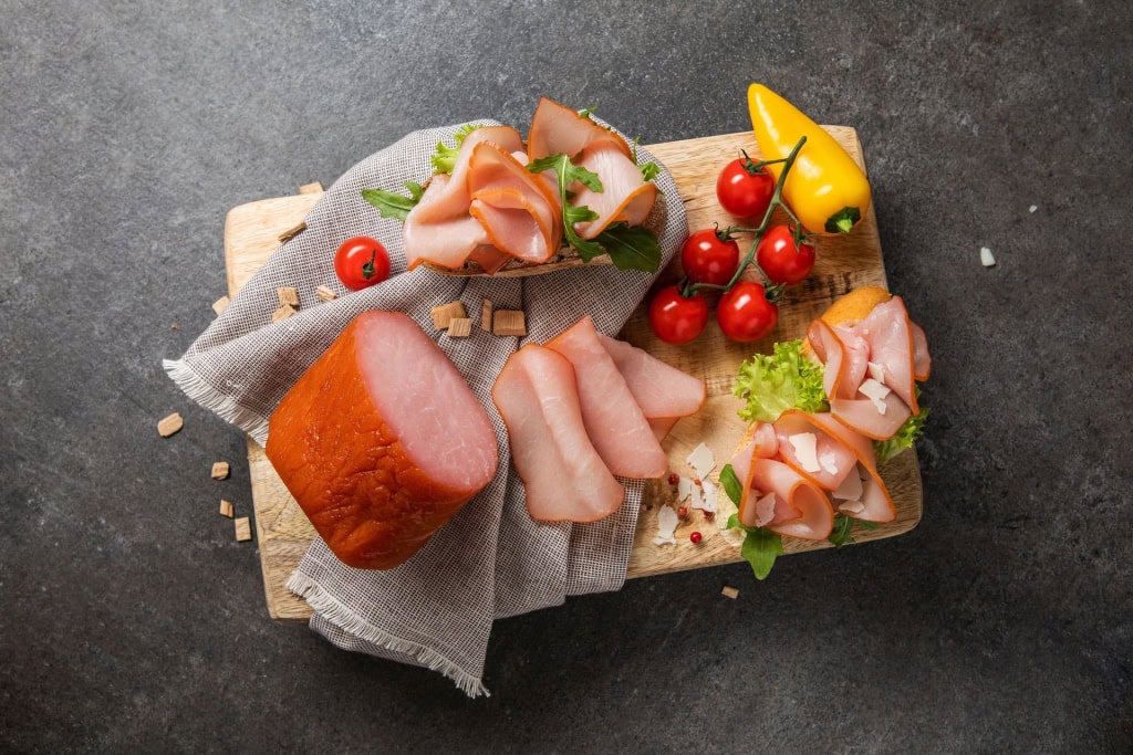 Lachsschinken in Scheiben und im Ganzen auf einem Holzbrett mit Kirschtomaten, Paprika, Salat und Schinkenbroten.